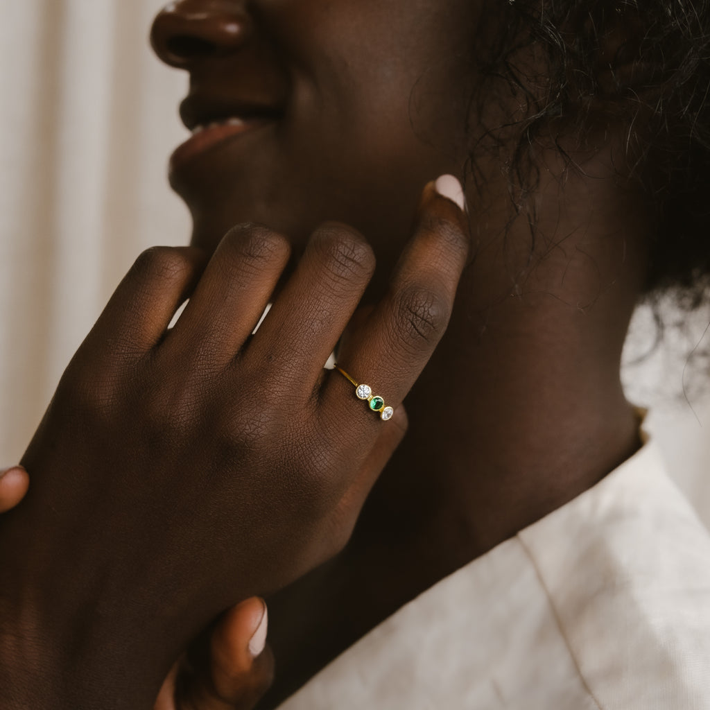 A woman smiles, displaying the Triple Birthstone Ring with green and clear stones on her finger.