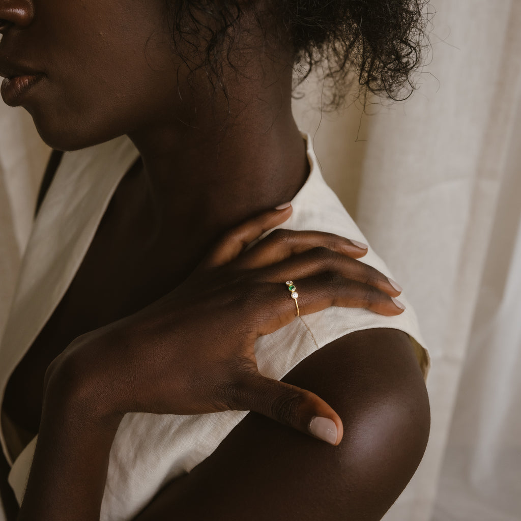 A woman in a cream top touches her shoulder, displaying the Triple Birthstone Ring with a green gemstone on her finger.