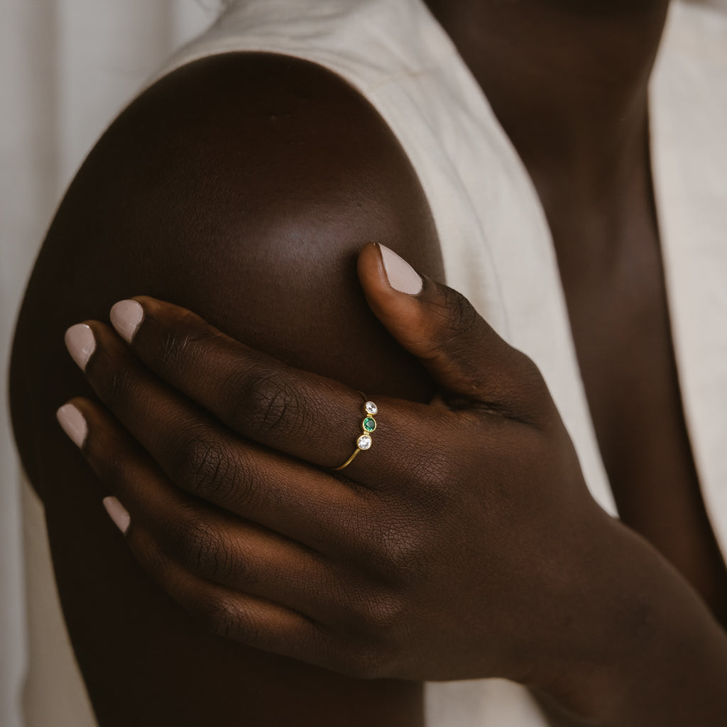 A person with dark skin wears the Triple Birthstone Ring, featuring three customizable gemstones, their hand resting gently on their shoulder.