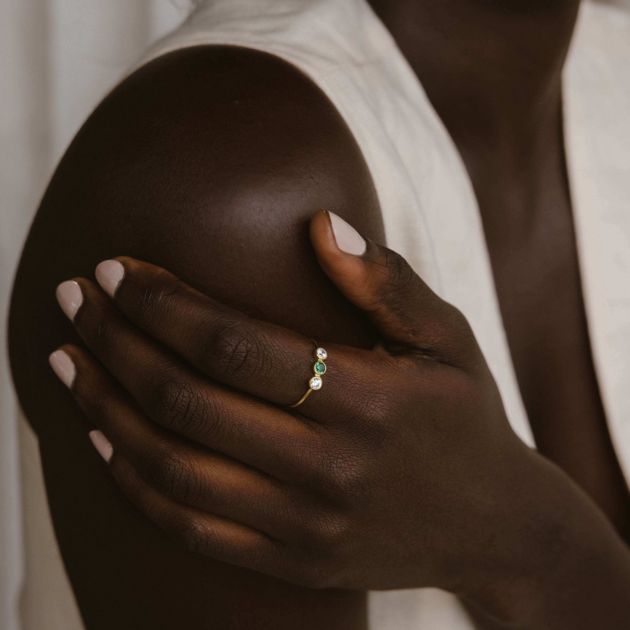 A person with dark skin wears the Triple Birthstone Ring, featuring three customizable gemstones, their hand resting gently on their shoulder.