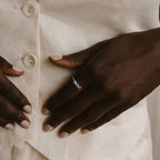 A hand adorned with the Triple Birthstone Ring, featuring customizable colorful stones, rests on a cream-colored button-up shirt.