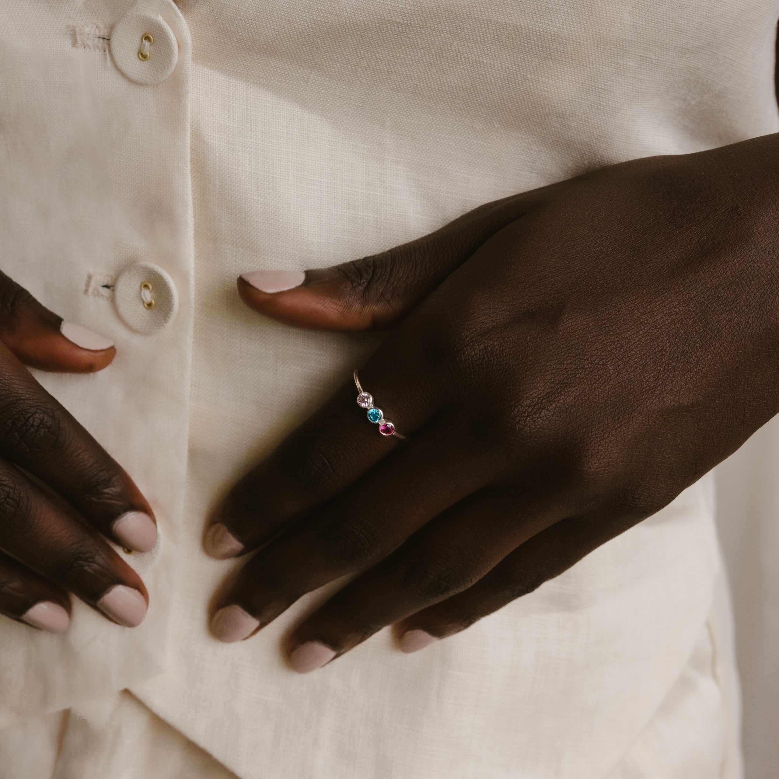 A hand adorned with the Triple Birthstone Ring, featuring customizable colorful stones, rests on a cream-colored button-up shirt.