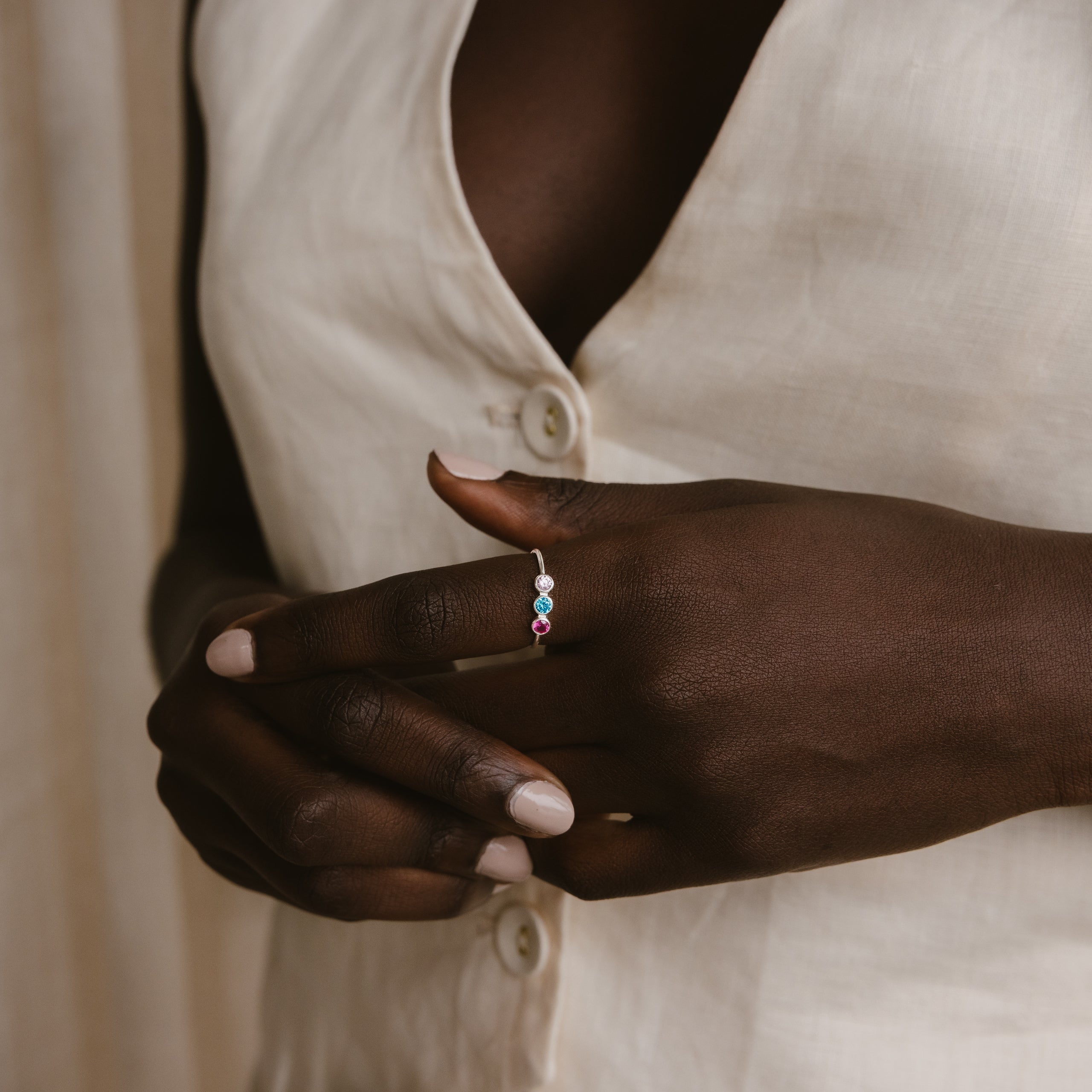 A person in a white buttoned shirt displays the Triple Birthstone Ring, featuring customizable colorful gemstones, on their finger.