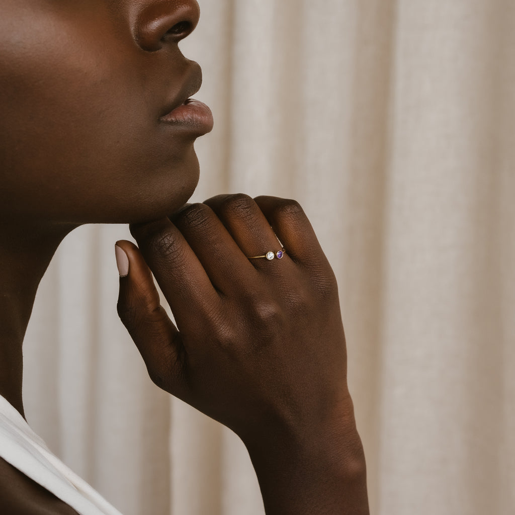 A woman with dark skin touches her chin, showcasing the Dual Birthstone Ring with a delicate pink stone on her finger.