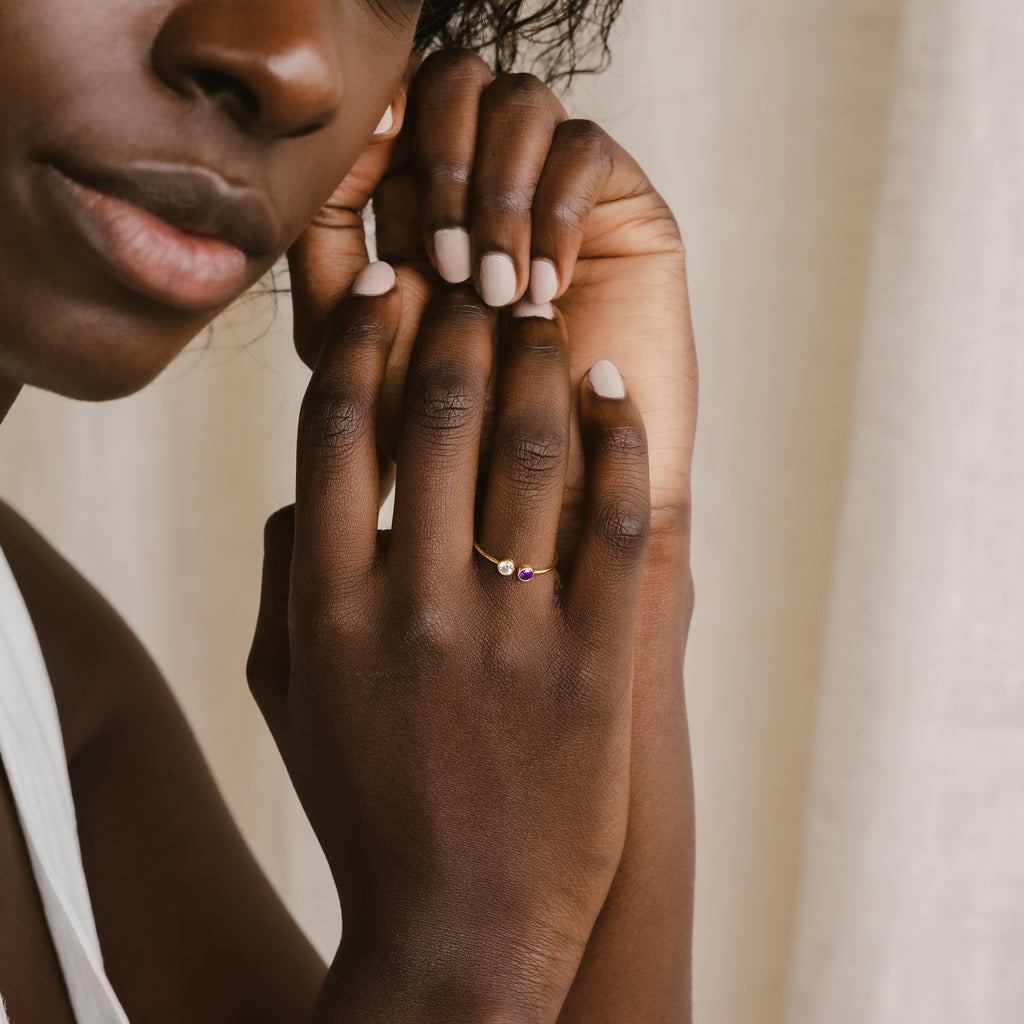 A woman with nude nails gently touches her face, wearing the Dual Birthstone Ring—a delicate wrap ring adorned with small purple gemstones.