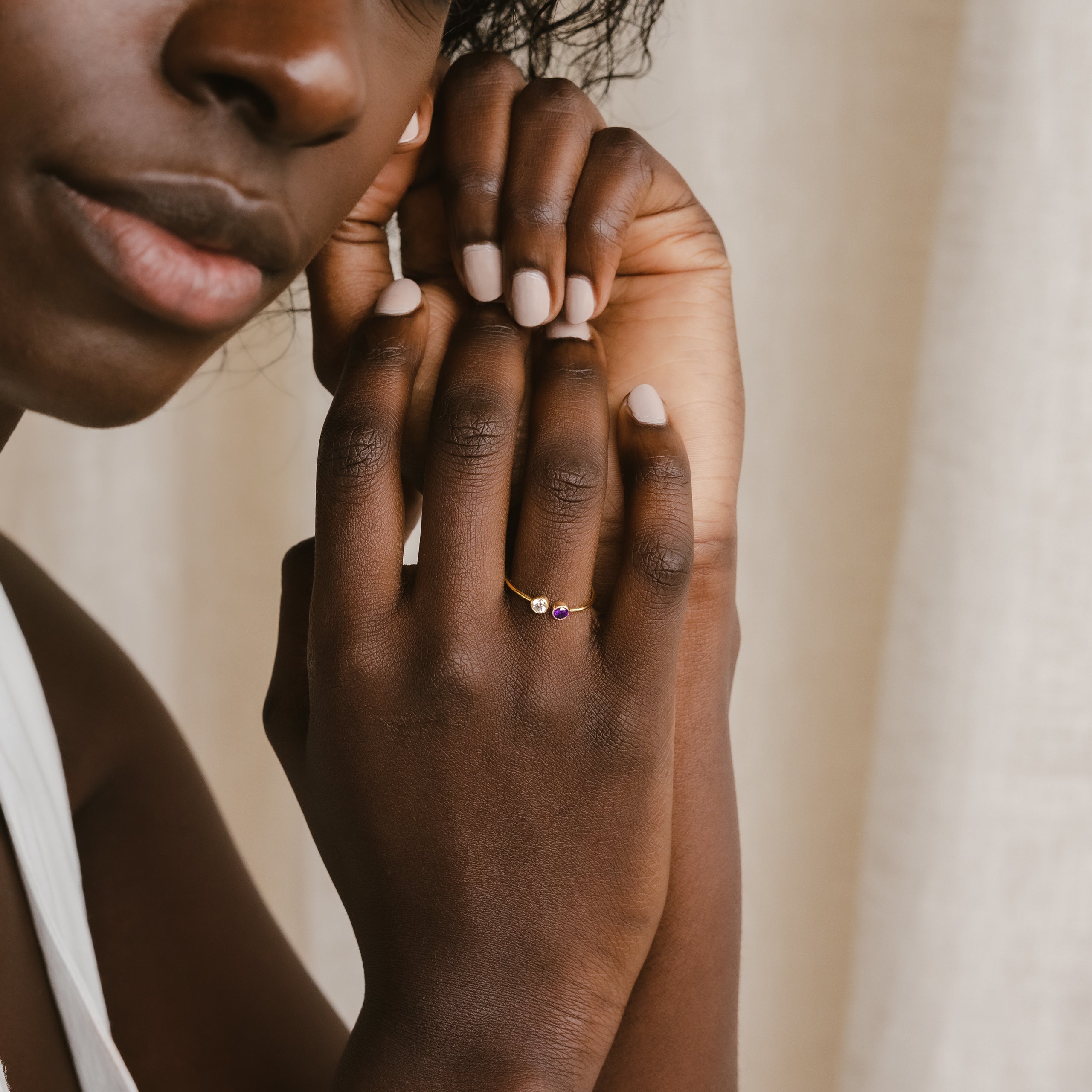 A woman with nude nails gently touches her face, wearing the Dual Birthstone Ring—a delicate wrap ring adorned with small purple gemstones.