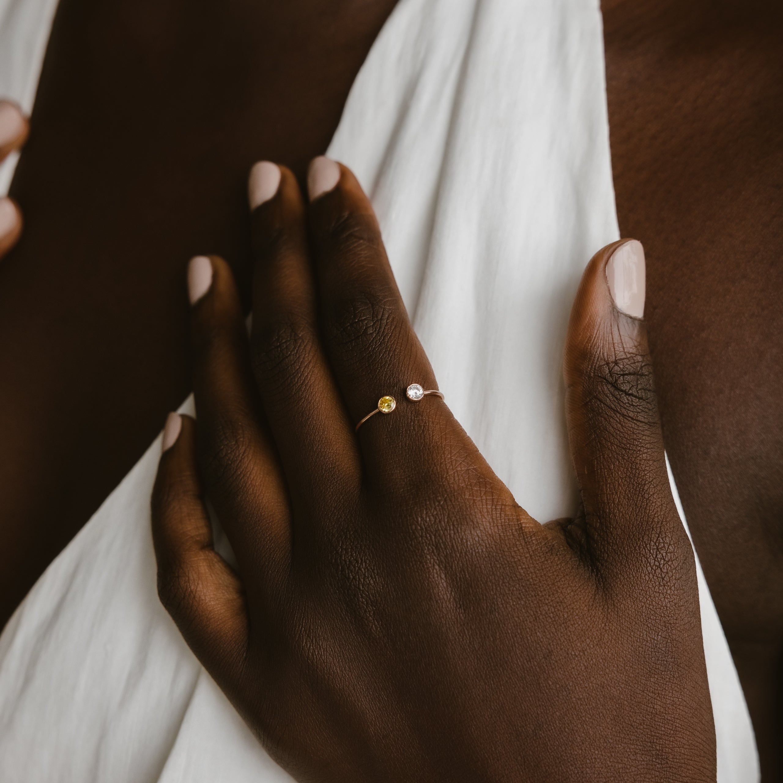 A hand with nude nails displays the Dual Birthstone Ring, a delicate wrap ring featuring two small stones, set against a white fabric background.