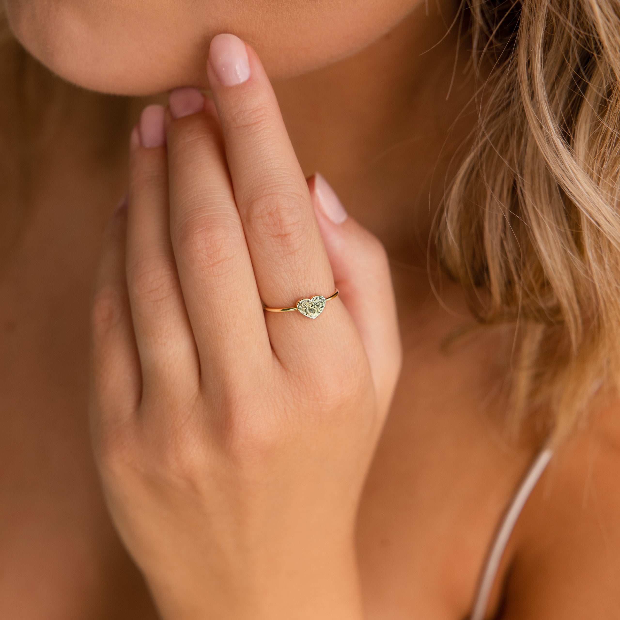 A woman's hand adorned with the Fingerprint Heart Ring touches her face; soft, natural lighting highlights this beautiful custom jewelry piece.