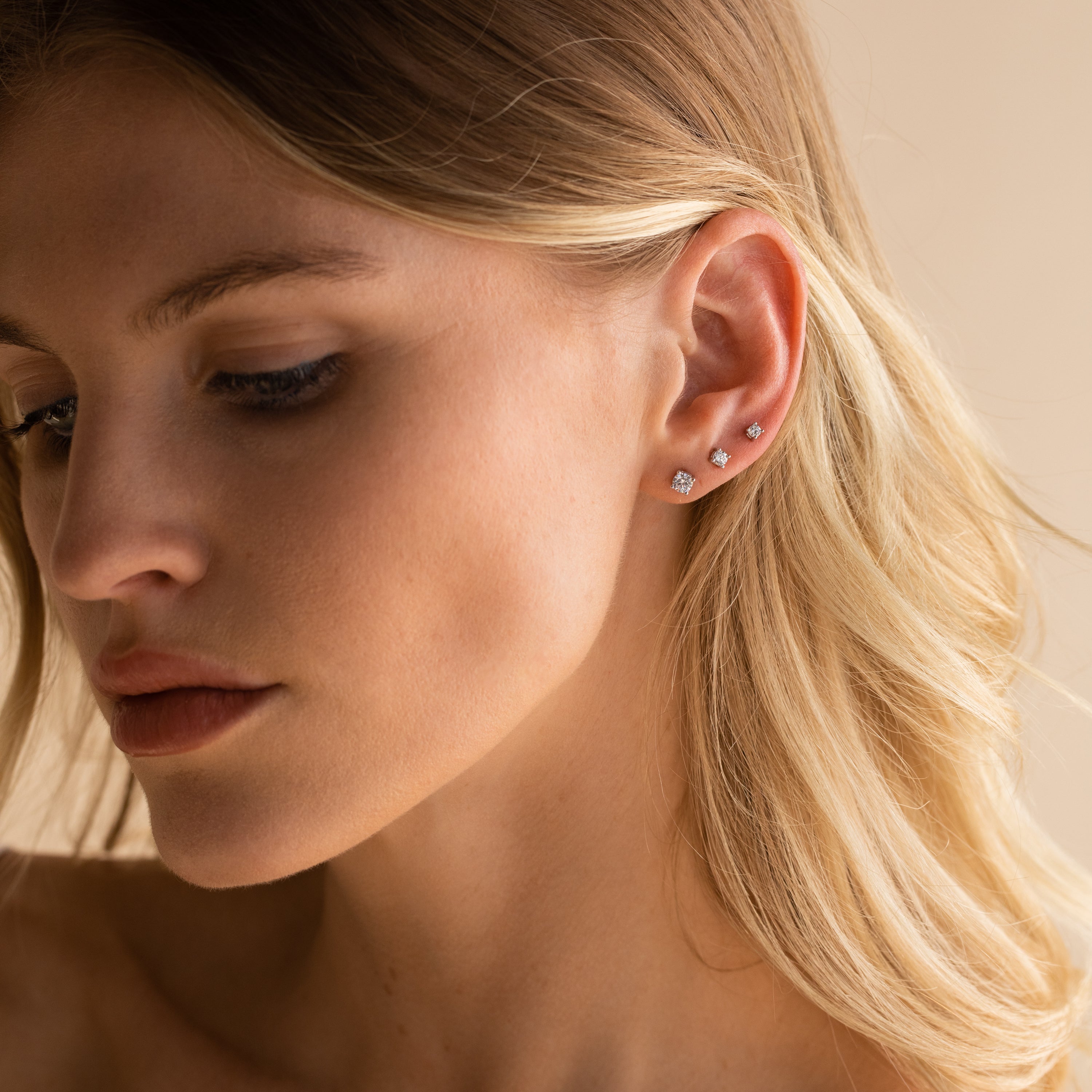 Woman with blonde hair wears three lightweight earrings, including the Booker Diamond Studs Set, on her left ear as she gazes down thoughtfully—an elegant and versatile choice for everyday jewelry.