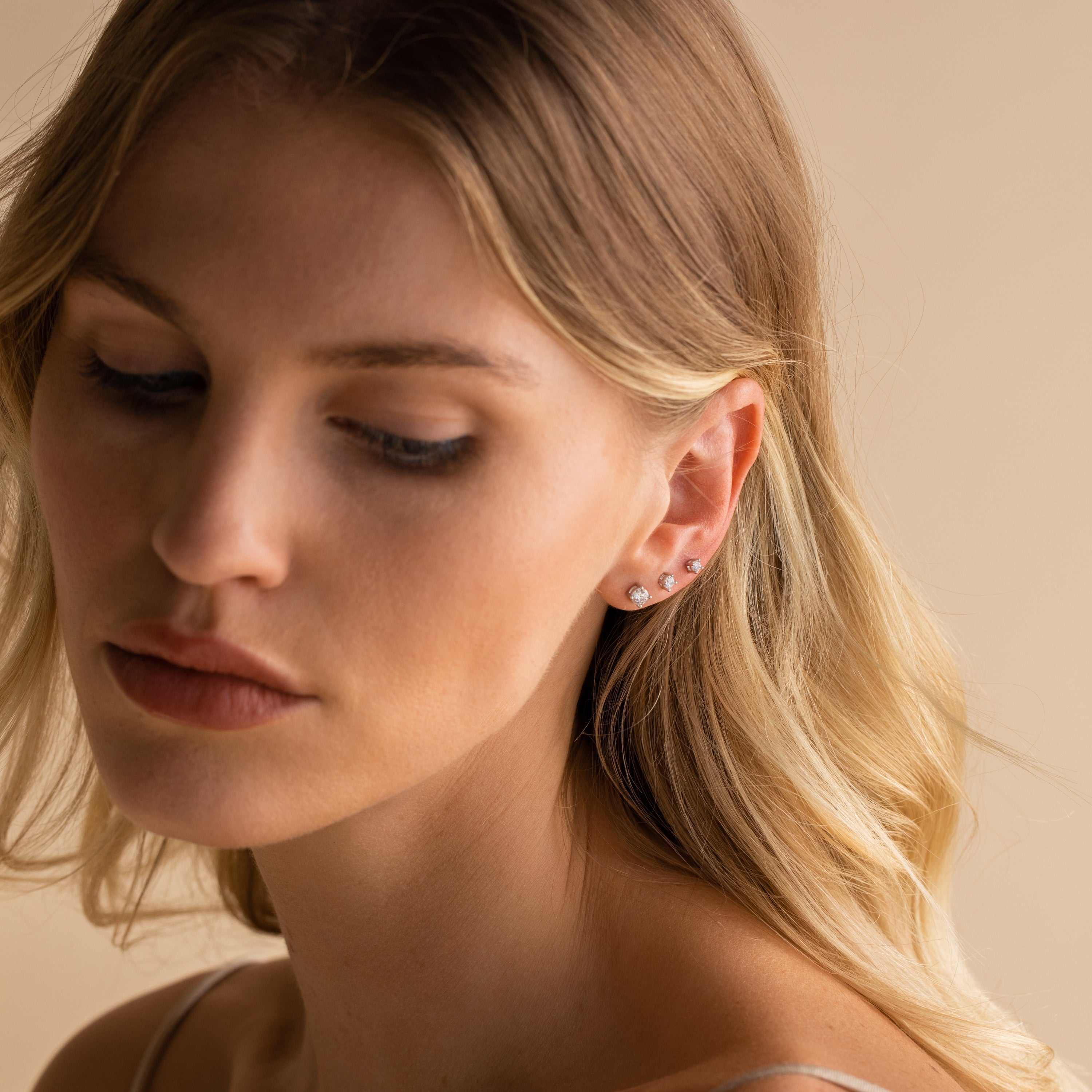 A woman with wavy blonde hair wears the Booker Diamond Studs Set—lightweight everyday earrings—while looking down against a beige background.