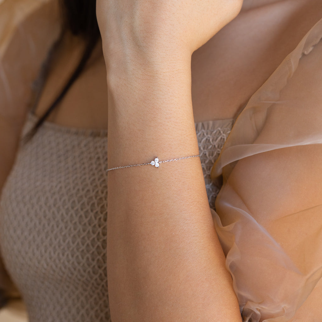 The Lily Diamond Bracelet, a delicate silver piece with a small charm, adorns a woman's wrist against a sheer beige sleeve—an elegant accessory for evening wear.