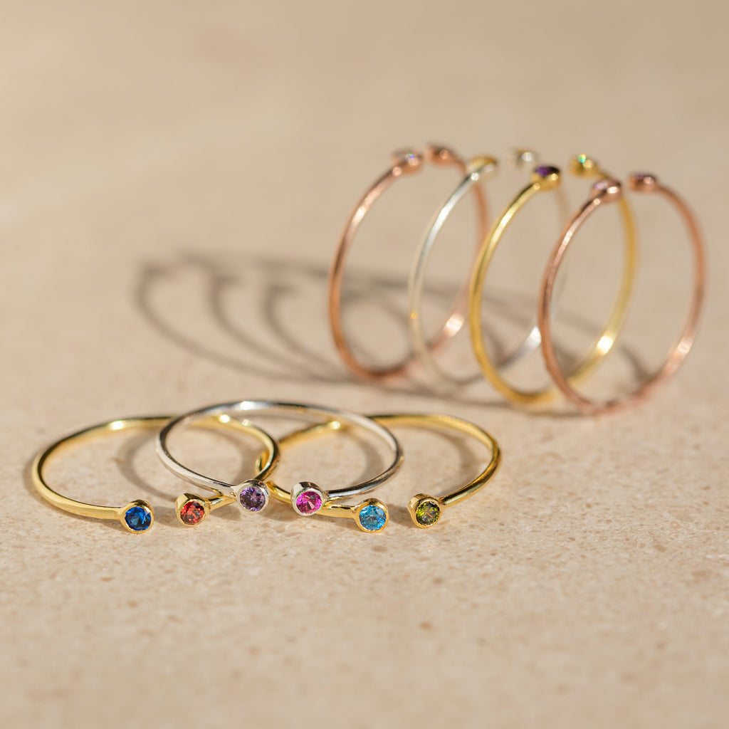 Five gold rings, such as the Tiny Dual Birthstone Ring with colorful gemstones, plus four thin hoop earrings are arranged on a beige surface.