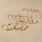 Five gold rings, such as the Tiny Dual Birthstone Ring with colorful gemstones, plus four thin hoop earrings are arranged on a beige surface.
