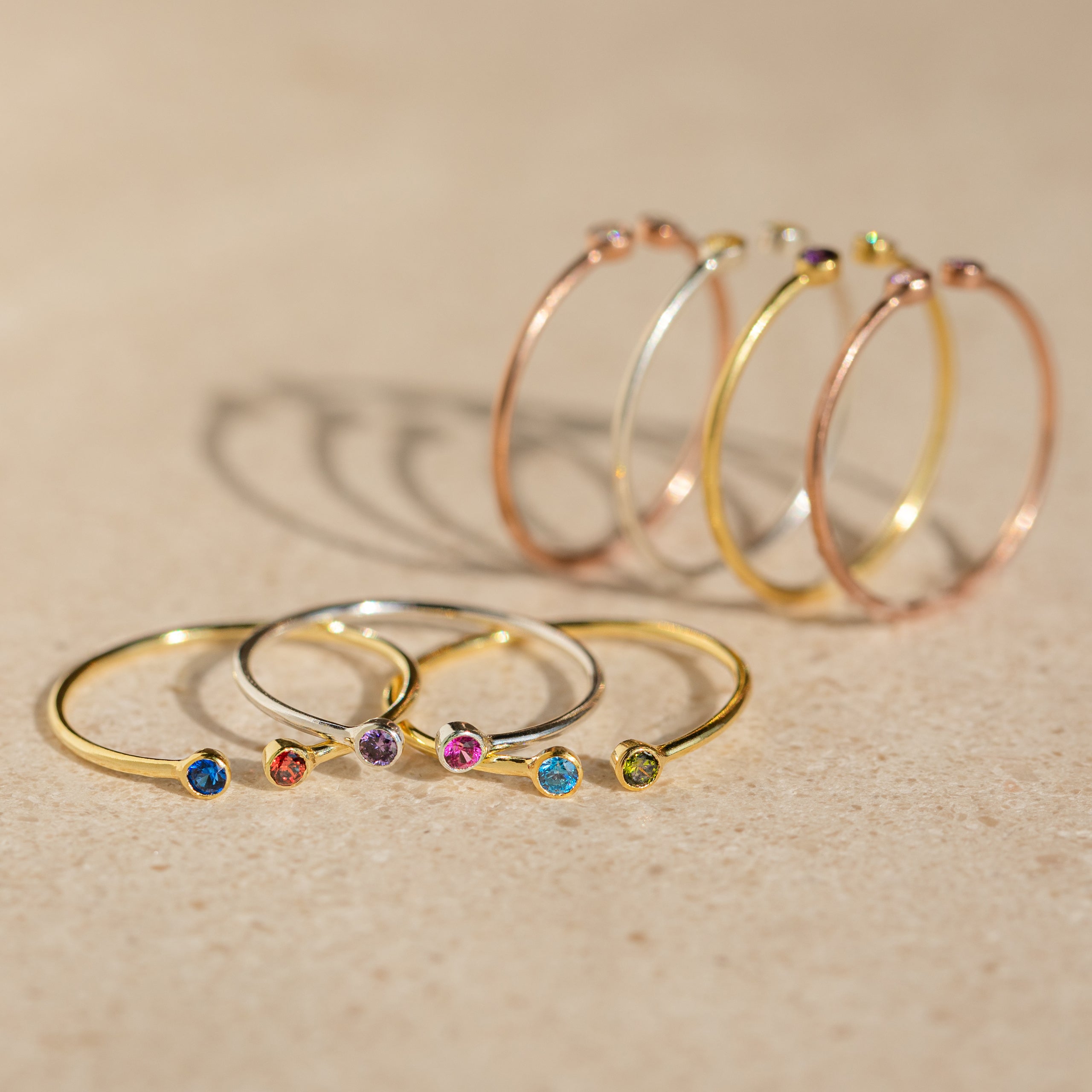 Five gold rings, such as the Tiny Dual Birthstone Ring with colorful gemstones, plus four thin hoop earrings are arranged on a beige surface.
