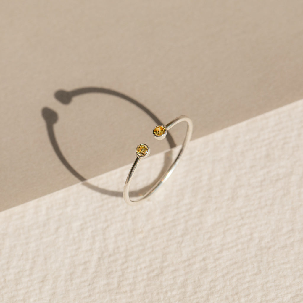 The Tiny Dual Birthstone Ring is a delicate silver open band adorned with two small yellow gems, casting subtle shadows on a textured two-toned surface.