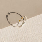 The Tiny Dual Birthstone Ring is a delicate silver open band adorned with two small yellow gems, casting subtle shadows on a textured two-toned surface.