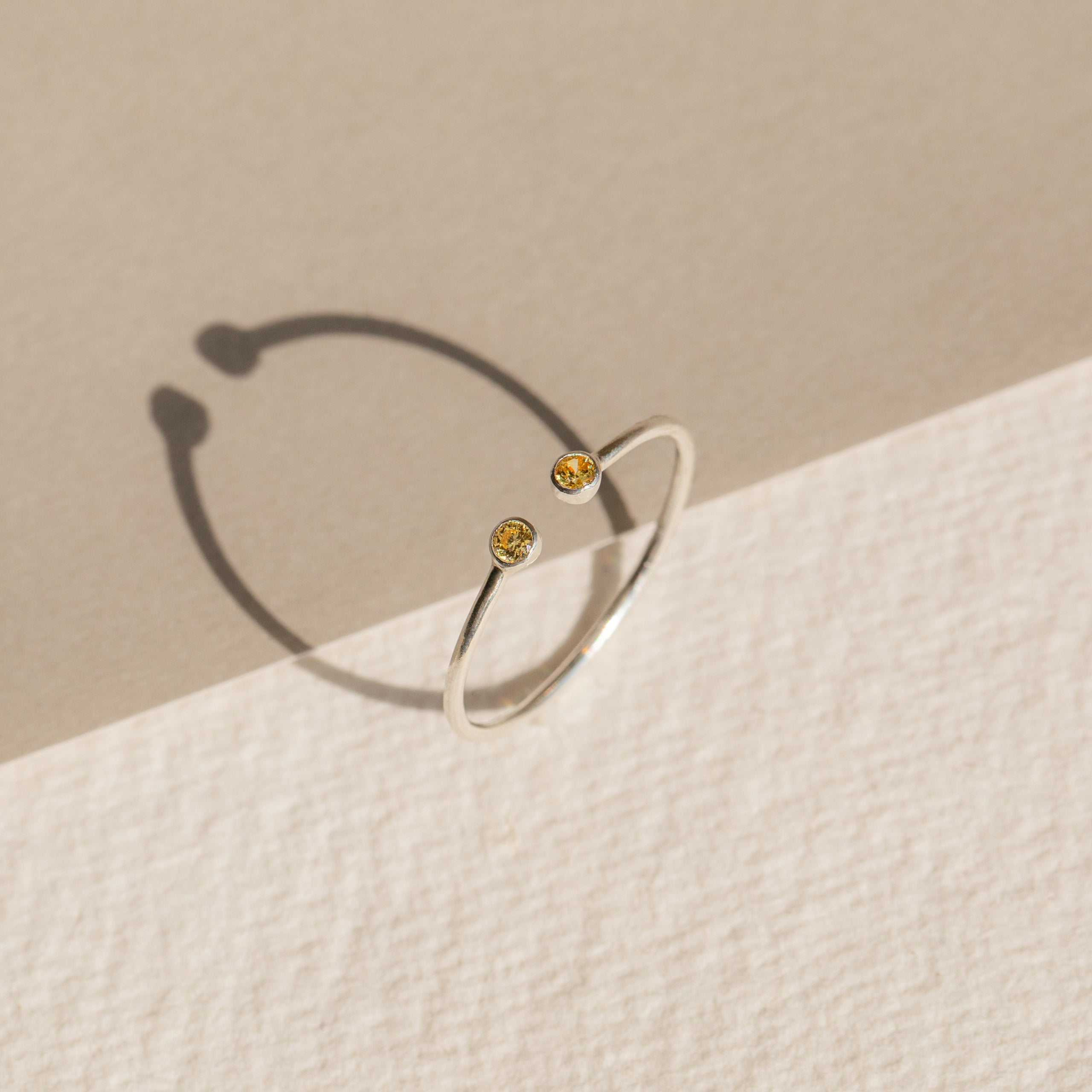 The Tiny Dual Birthstone Ring is a delicate silver open band adorned with two small yellow gems, casting subtle shadows on a textured two-toned surface.