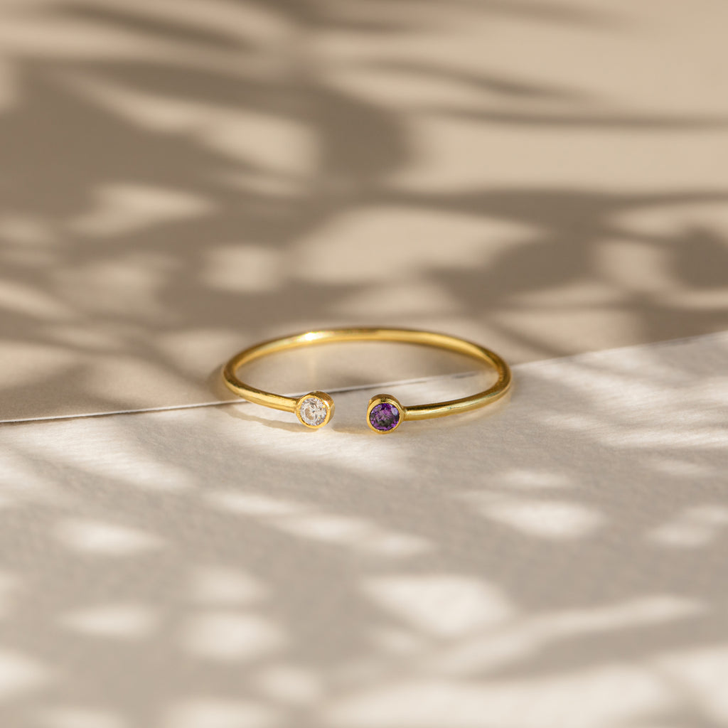 The Tiny Dual Birthstone Ring showcases clear and purple stones on a textured surface with soft shadows—an elegant and dainty ring, perfect for daily wear.