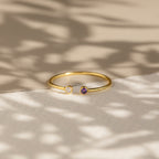 The Tiny Dual Birthstone Ring showcases clear and purple stones on a textured surface with soft shadows—an elegant and dainty ring, perfect for daily wear.