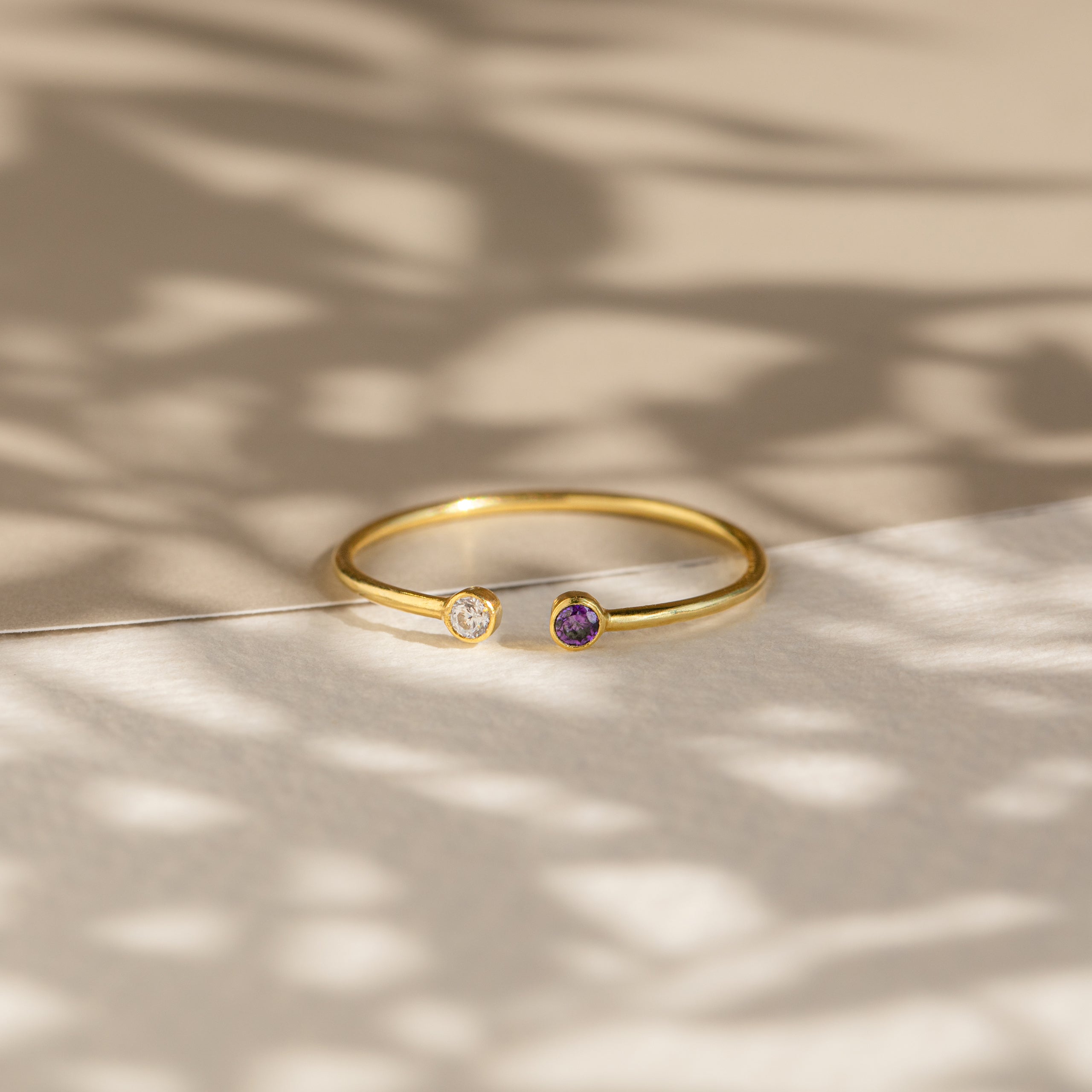 The Tiny Dual Birthstone Ring showcases clear and purple stones on a textured surface with soft shadows—an elegant and dainty ring, perfect for daily wear.