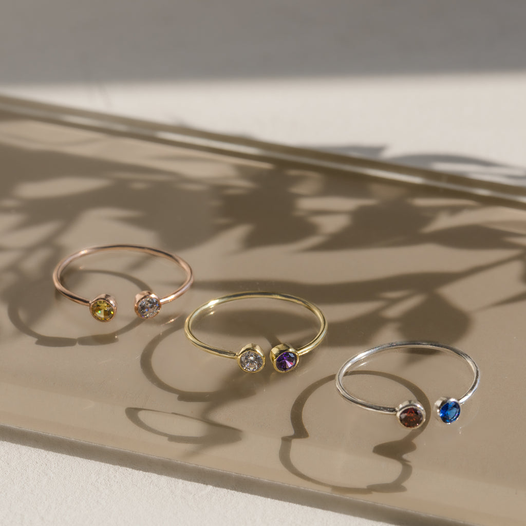 The Dual Birthstone Ring, a delicate wrap ring featuring colorful gemstones, rests on a reflective surface surrounded by soft shadows and sunlight.
