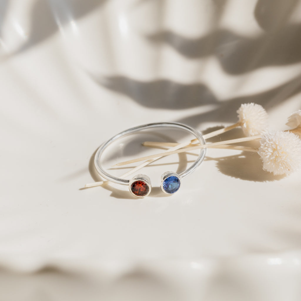 This Dual Birthstone Ring, featuring red and blue gemstones, is shown beside dried white flowers on a silver shell-shaped dish.