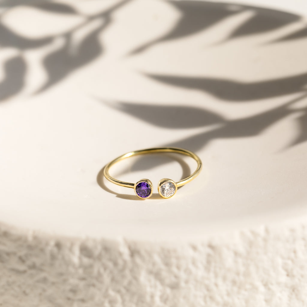 The Dual Birthstone Ring is a delicate gold wrap ring with one purple and one clear gemstone, displayed elegantly on a white textured surface with leafy shadows.
