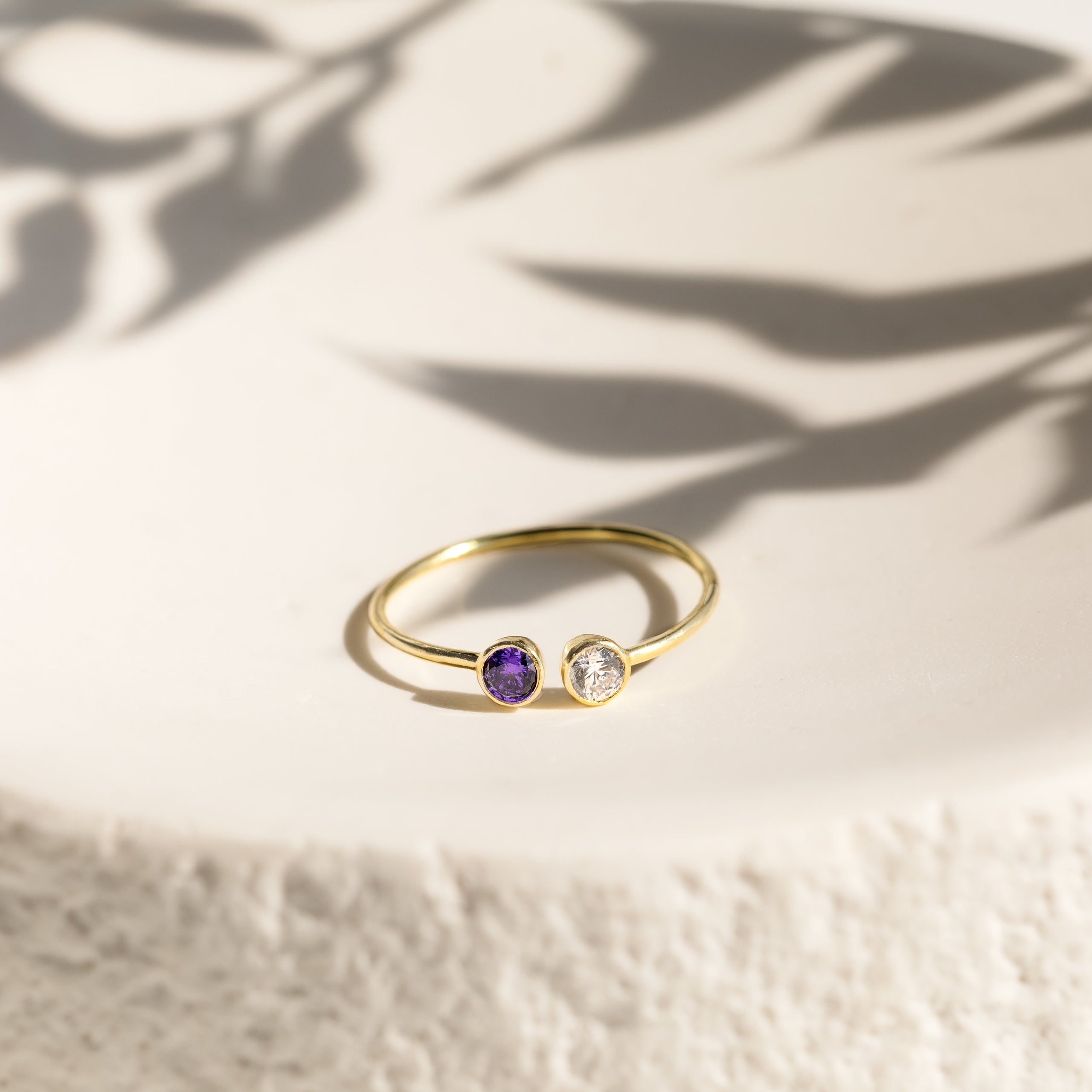 The Dual Birthstone Ring is a delicate gold wrap ring with one purple and one clear gemstone, displayed elegantly on a white textured surface with leafy shadows.
