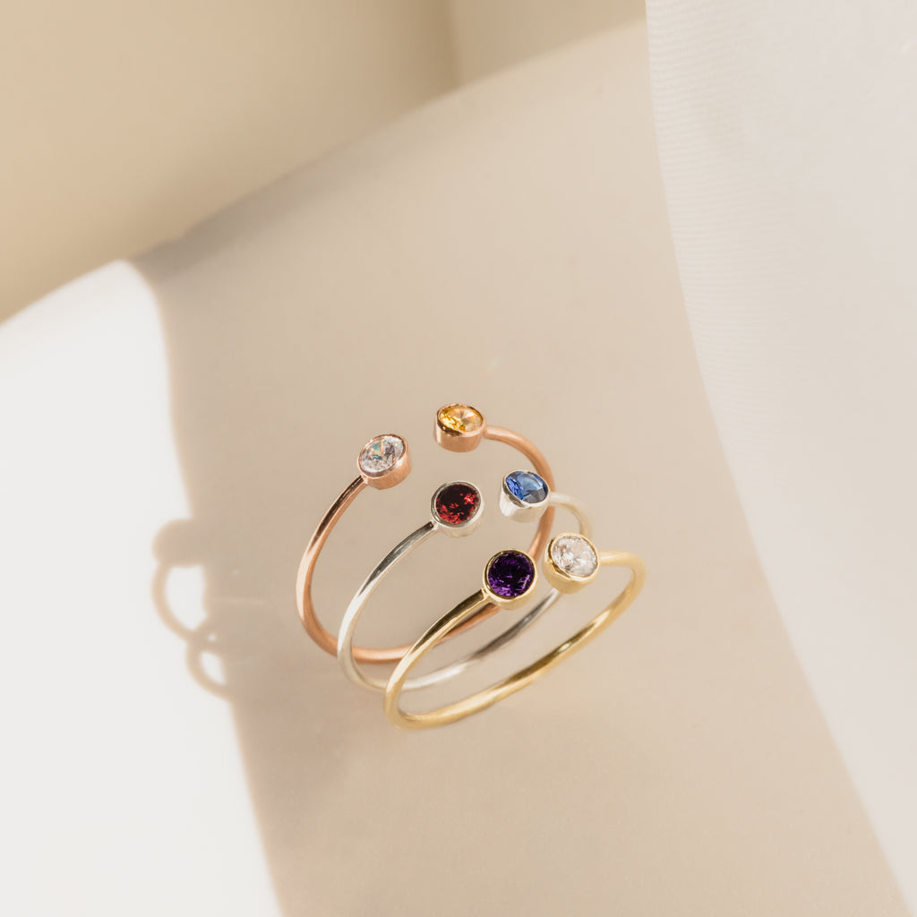 The Dual Birthstone Ring, shown with three other delicate rings, features colorful gemstones and is displayed on a light surface in soft natural light.