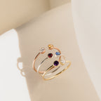 The Dual Birthstone Ring, shown with three other delicate rings, features colorful gemstones and is displayed on a light surface in soft natural light.