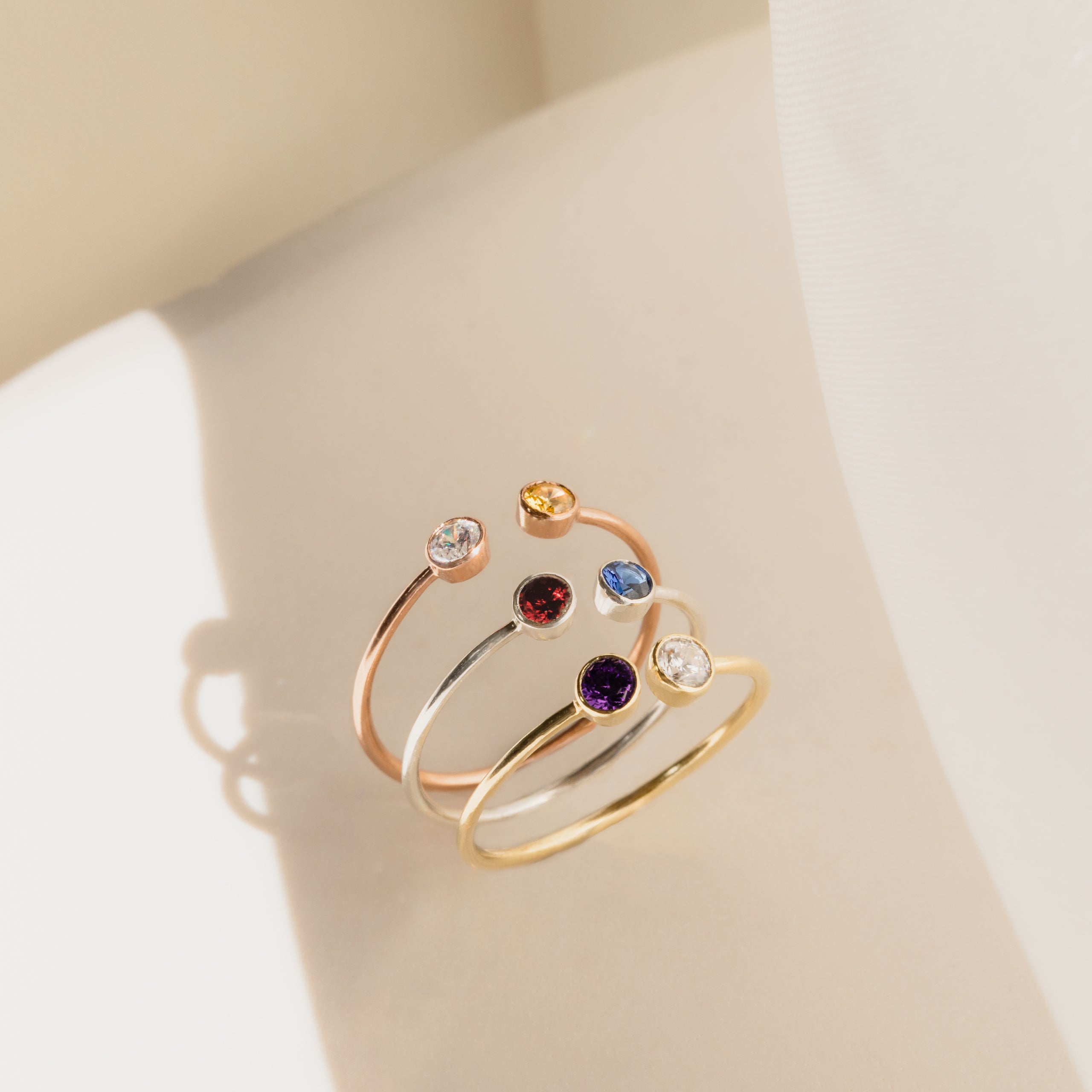 The Dual Birthstone Ring, shown with three other delicate rings, features colorful gemstones and is displayed on a light surface in soft natural light.