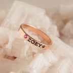 The Stacking Name Band is a rose gold stackable ring engraved with ZOEY and a paw print, featuring a small pink gemstone set on white crystals.