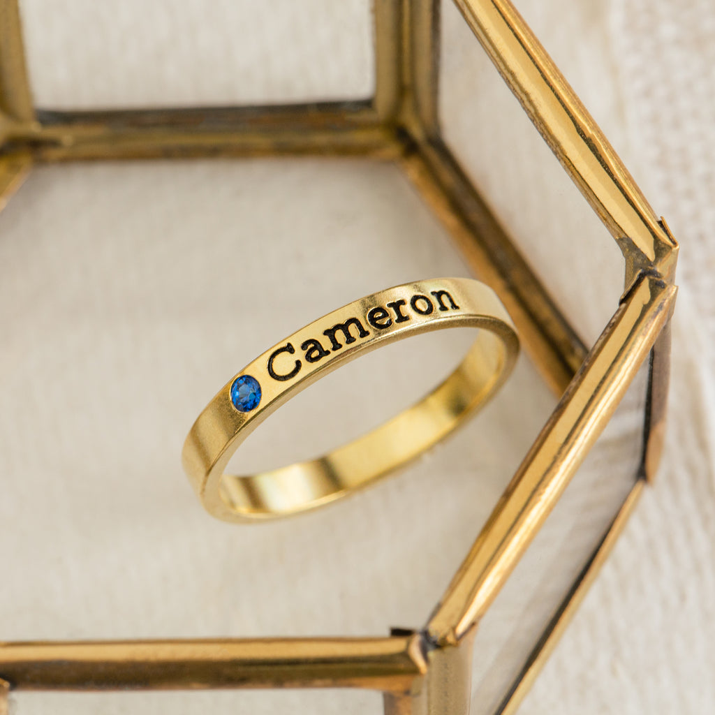 The Mothers Birthstone Ring, engraved with the name Cameron and featuring a delicate blue gemstone, creates an elegant personalized keepsake when displayed on a glass surface.