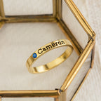 The Mothers Birthstone Ring, engraved with the name Cameron and featuring a delicate blue gemstone, creates an elegant personalized keepsake when displayed on a glass surface.