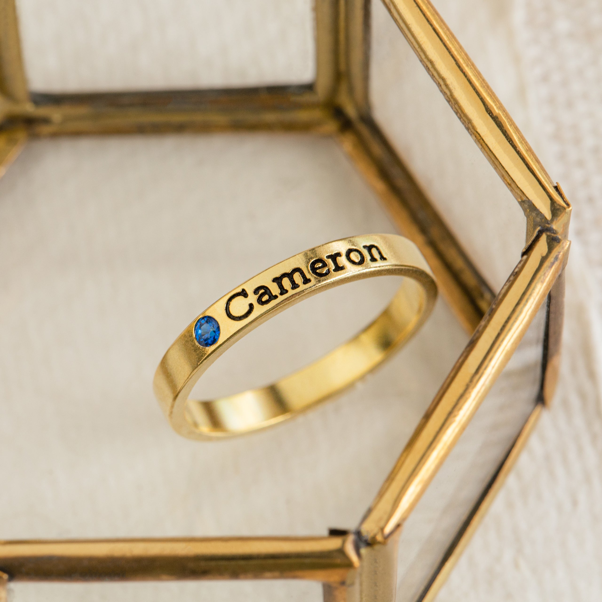 The Mothers Birthstone Ring, engraved with the name Cameron and featuring a delicate blue gemstone, creates an elegant personalized keepsake when displayed on a glass surface.