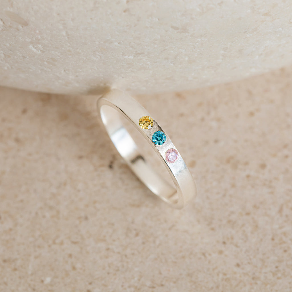 Mothers Birthstone Ring with customizable yellow, blue, and pink gemstones set in a row, displayed on a light beige stone surface.