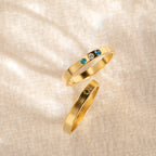 Two gold rings lie on textured fabric: one is the Mothers Birthstone Ring with three customizable gemstones for a personal touch, while the other remains plain.