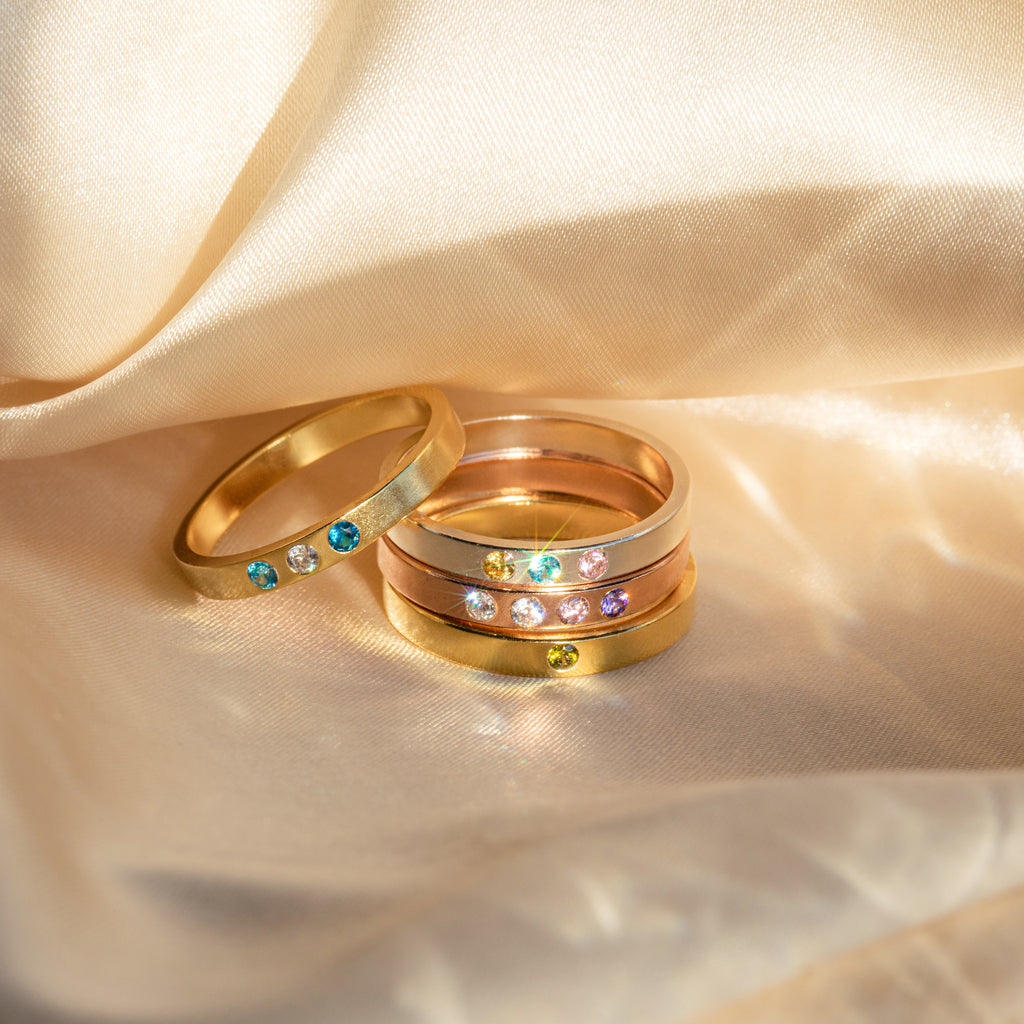 The Mothers Birthstone Ring features customizable gemstones set in gold and silver, displayed on champagne-colored satin in sunlight for a unique personalized keepsake.