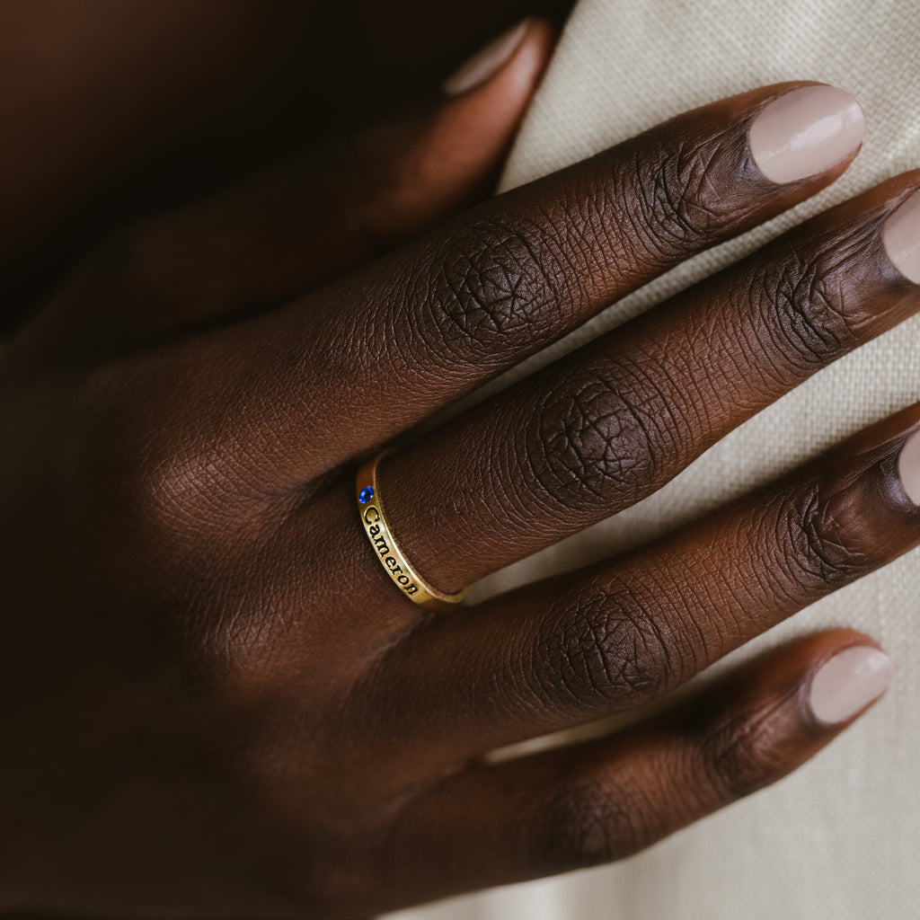 A hand with neatly manicured nails wears the Mothers Birthstone Ring, a gold band engraved with Cancer and set with a small blue gemstone—an ideal personalized keepsake.