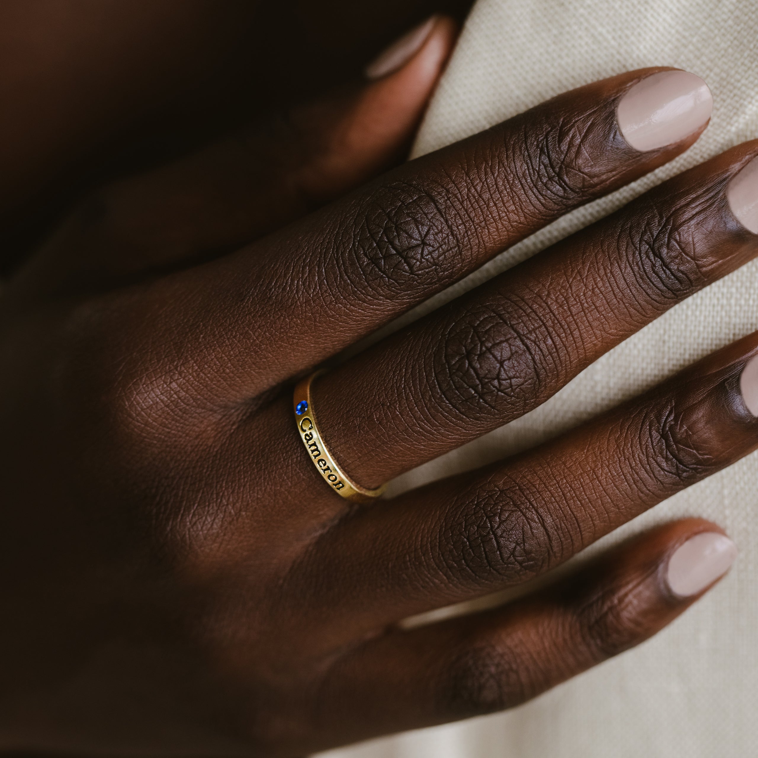 A hand with neatly manicured nails wears the Mothers Birthstone Ring, a gold band engraved with Cancer and set with a small blue gemstone—an ideal personalized keepsake.