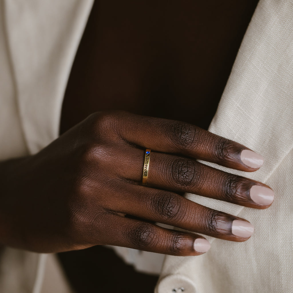 A hand with pale pink nails wears the Stacking Name Band—gold with a small blue stone—resting on cream-colored fabric.