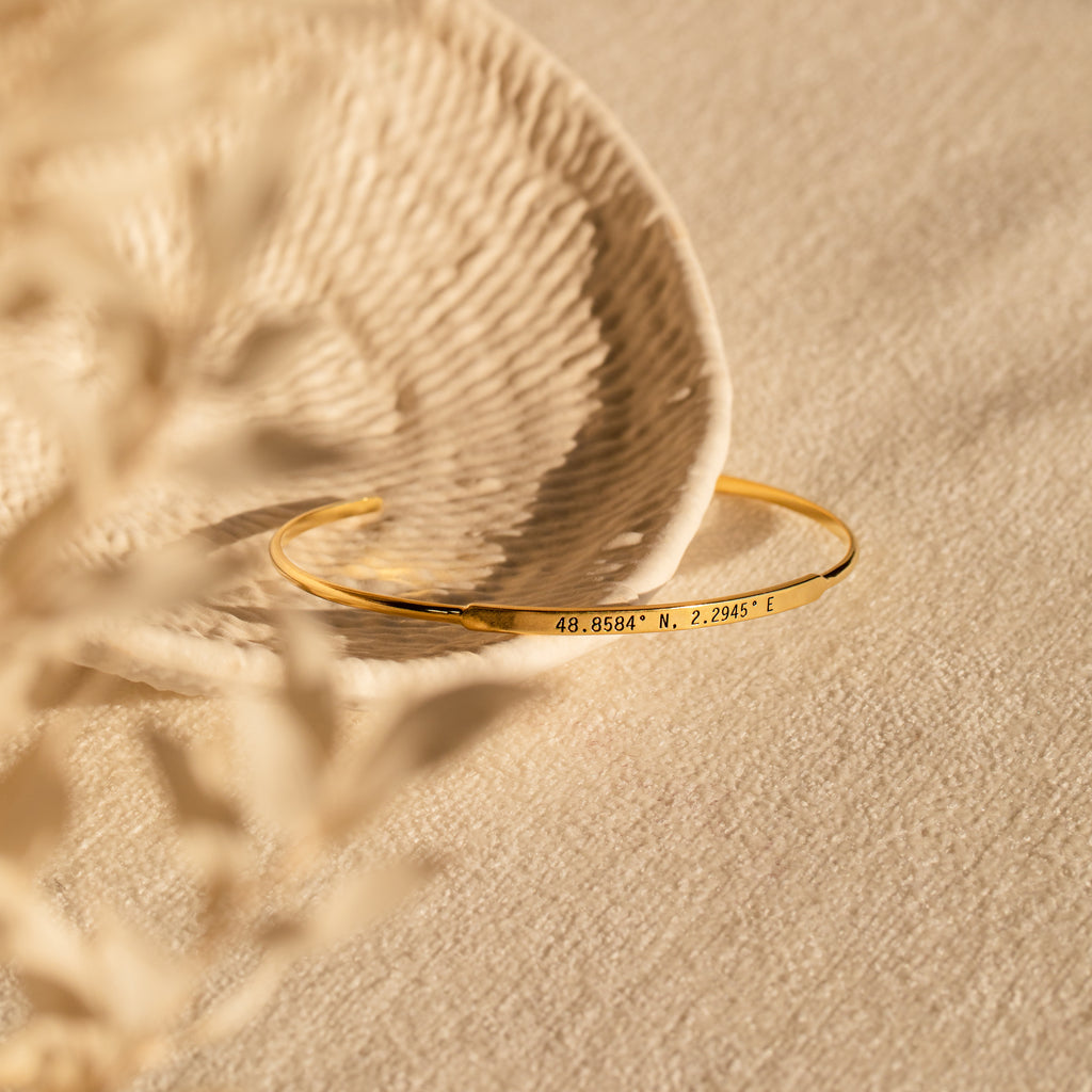 The Custom Message Cuff, a gold engraved bracelet with coordinates, rests on a textured cream surface near a decorative dish—a unique personalized gift for someone special.