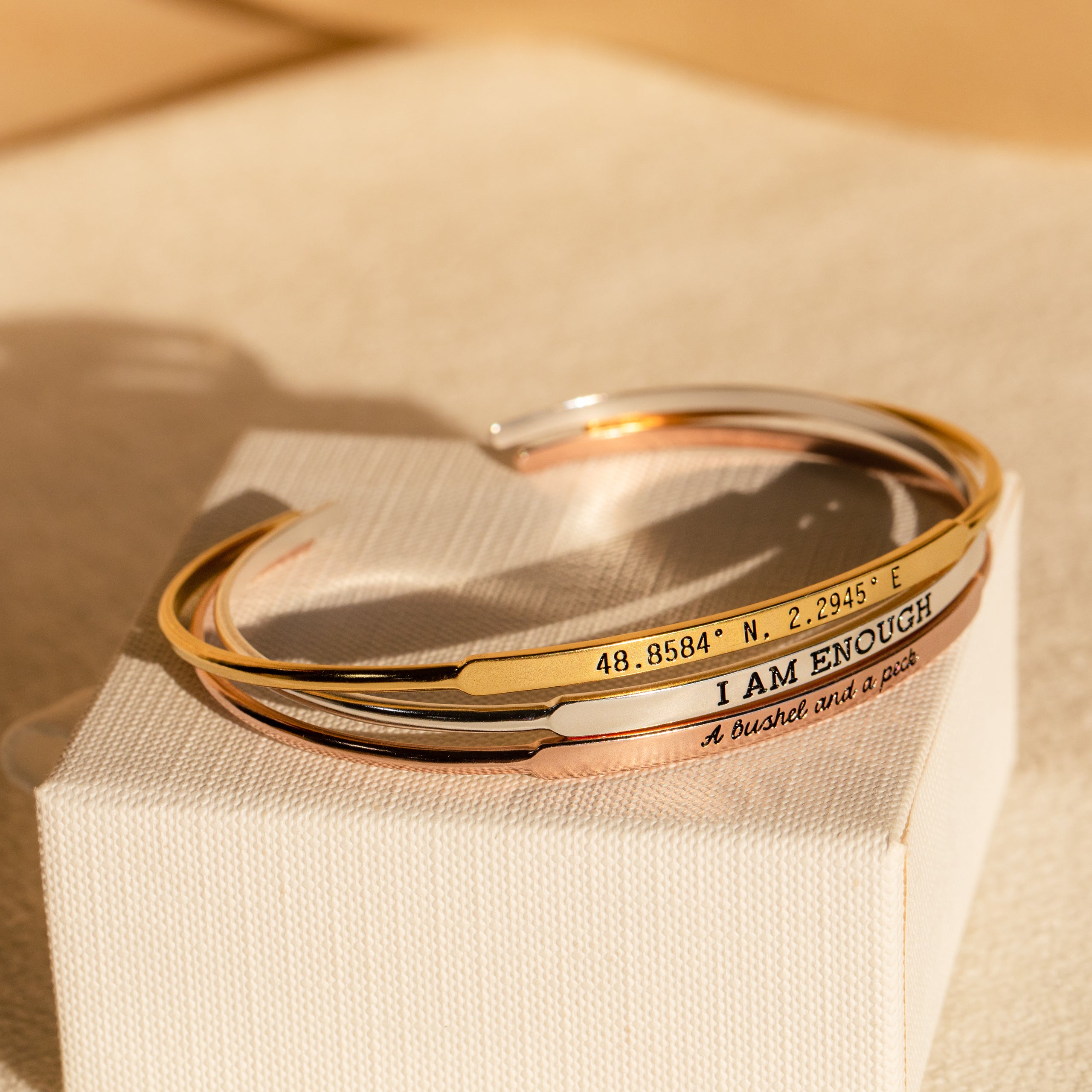 Three Custom Message Cuff bracelets with inspiring engravings—like I AM ENOUGH—rest on a textured cream box, making an elegant and personalized gift for any occasion.