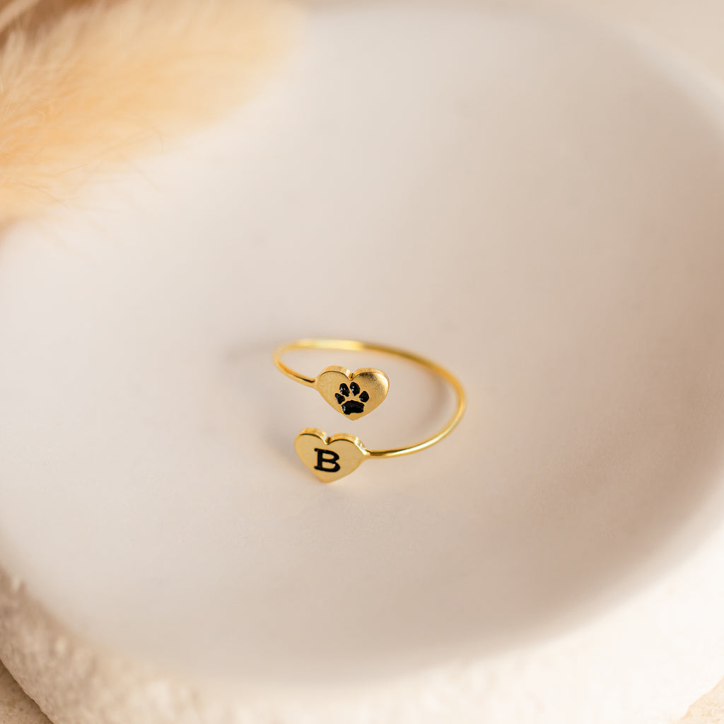 The Paw Print Duo Heart Ring features two gold hearts—one with a black paw print and the other with the letter B—shown on a white dish. A charming Pet Lovers Gift, perfect for showcasing your love for pets.