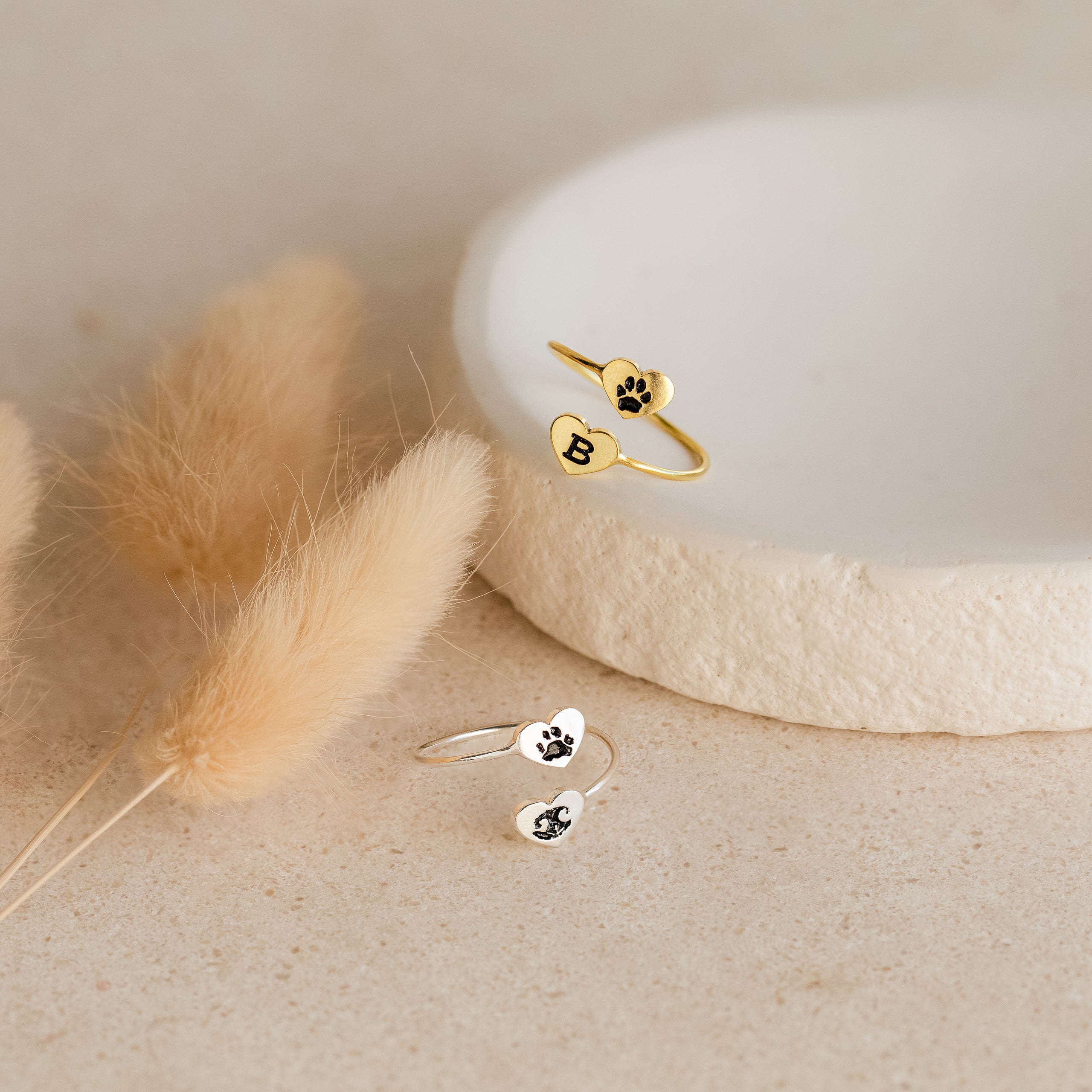 The Paw Print Duo Heart Ring rests on a stone dish beside fluffy beige dried flowers, making it the perfect gift for pet lovers.