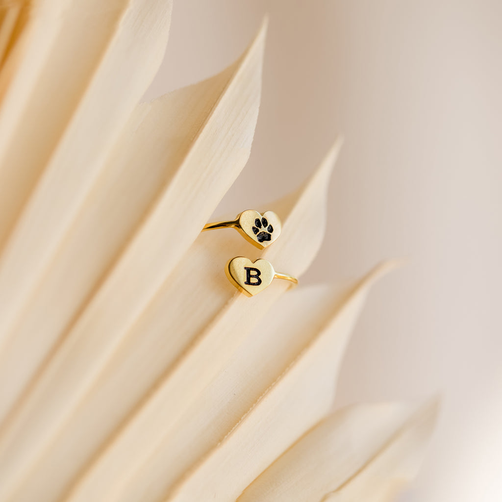 The Paw Print Duo Heart Ring features heart-shaped ends—one with a paw print, the other with a "B"—resting on beige leaves. A perfect Pet Lovers Gift to honor your furry friend.