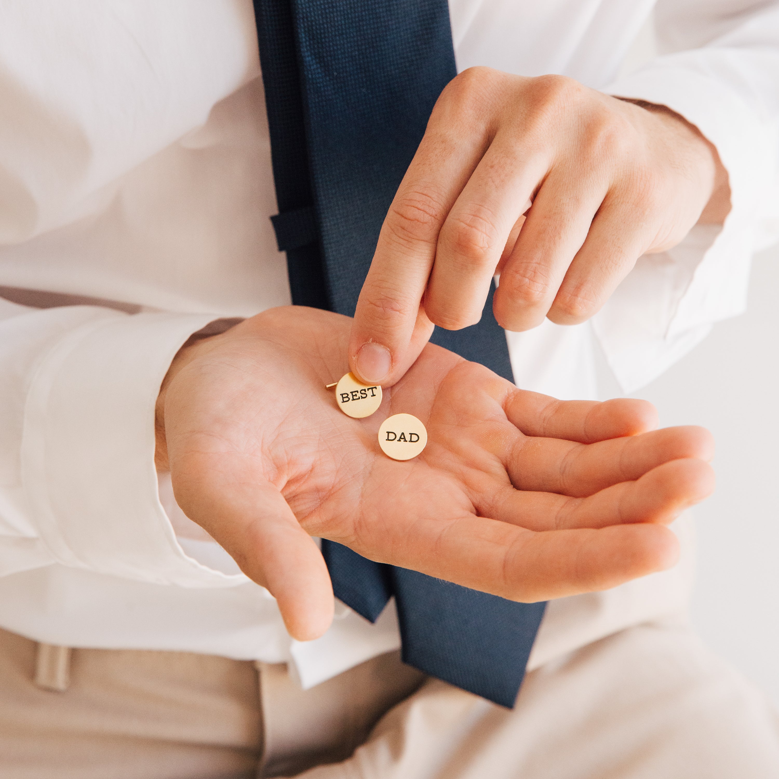 A man in a tie holds two round pins reading BEST and DAD, resembling the Engraved Cufflinks—an ideal personalized gift for celebrating fatherhood.