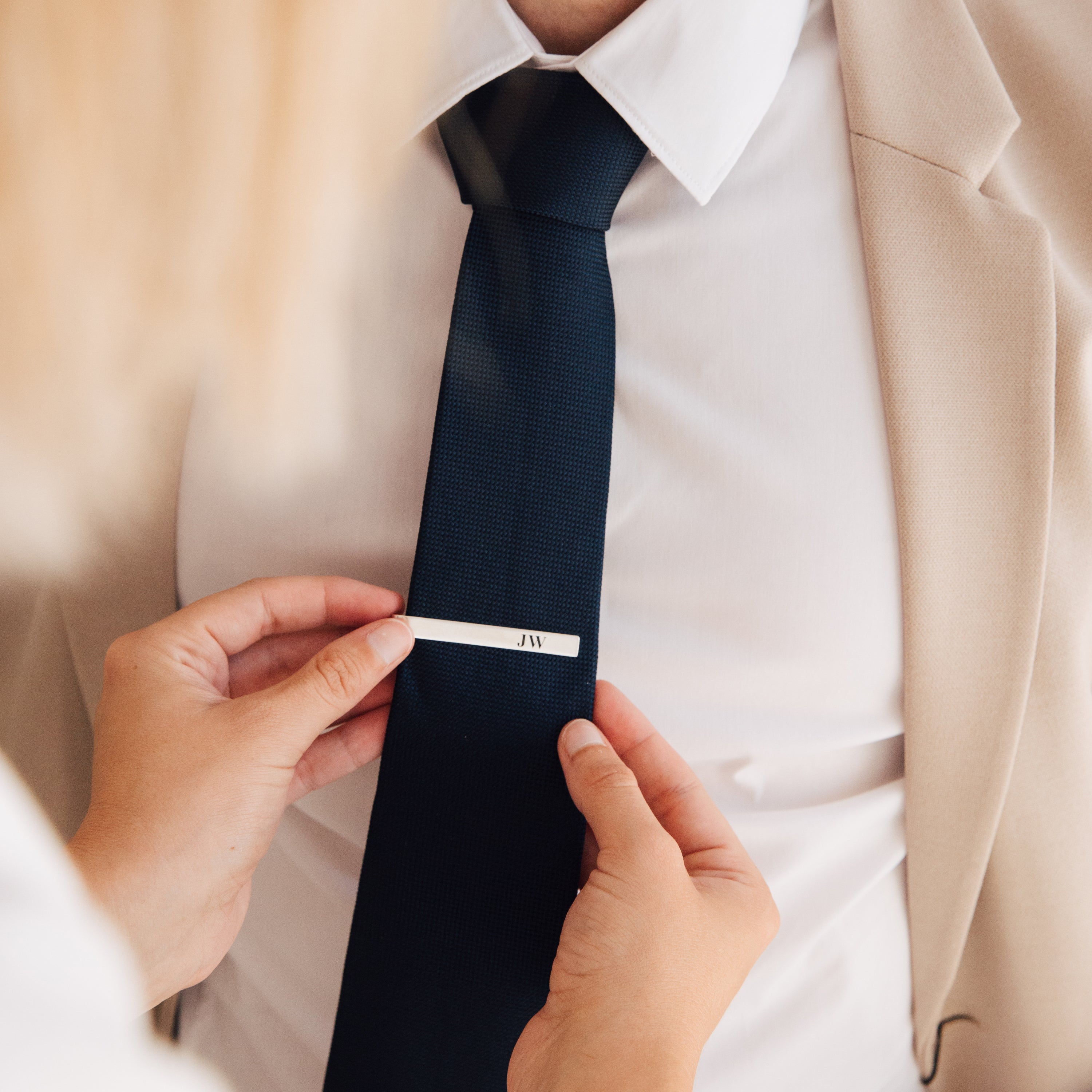 A person adjusts a navy tie with the Handwriting Tie Clip on someone dressed in a white shirt and beige blazer.