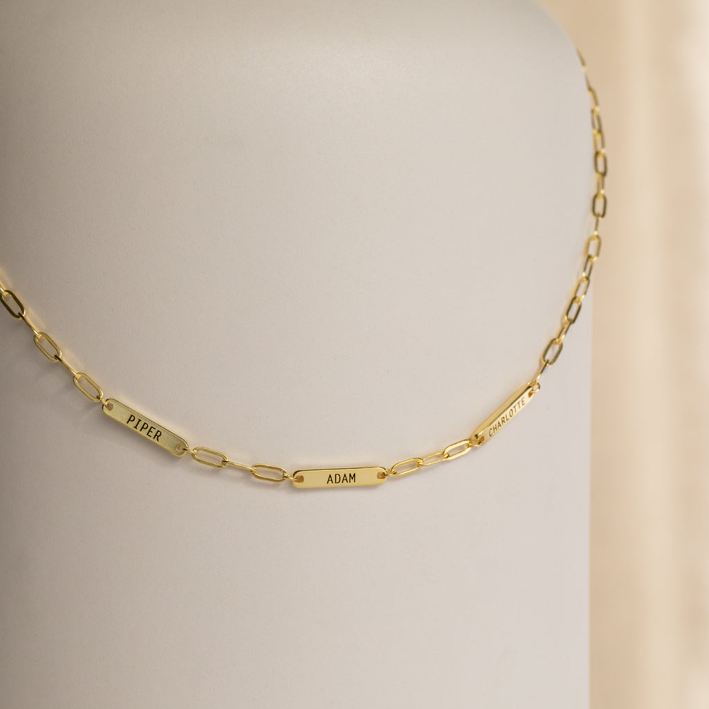 The Mini Name Bar Necklace showcases a gold chain with three custom name tags—Piper, Adam, and Charlotte—set against a neutral background.