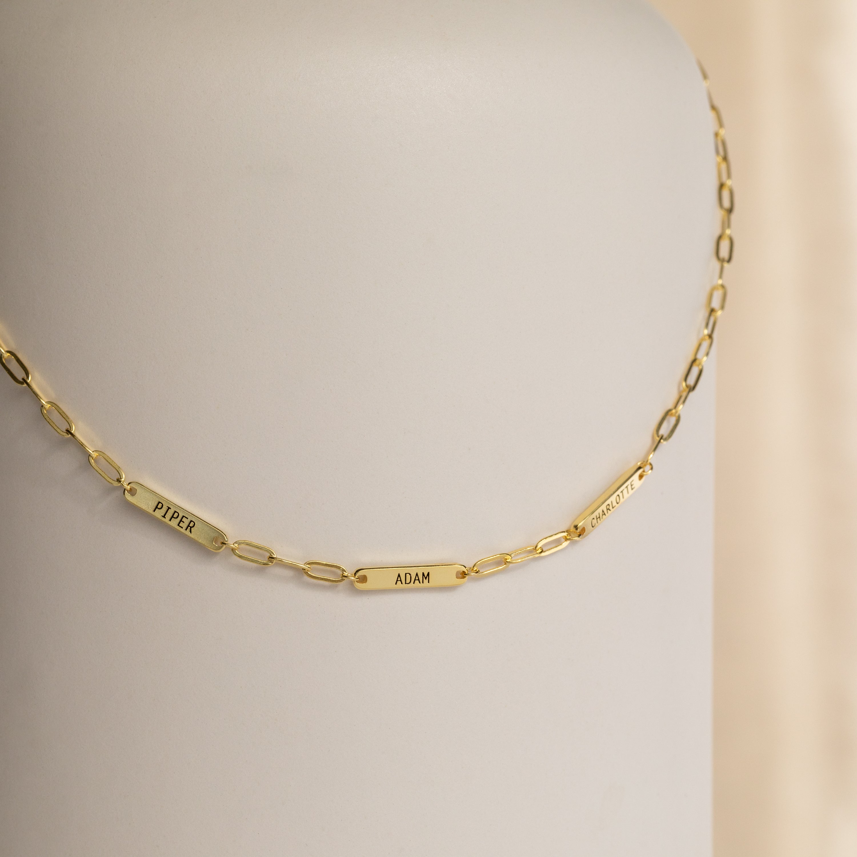 The Mini Name Bar Necklace showcases a gold chain with three custom name tags—Piper, Adam, and Charlotte—set against a neutral background.